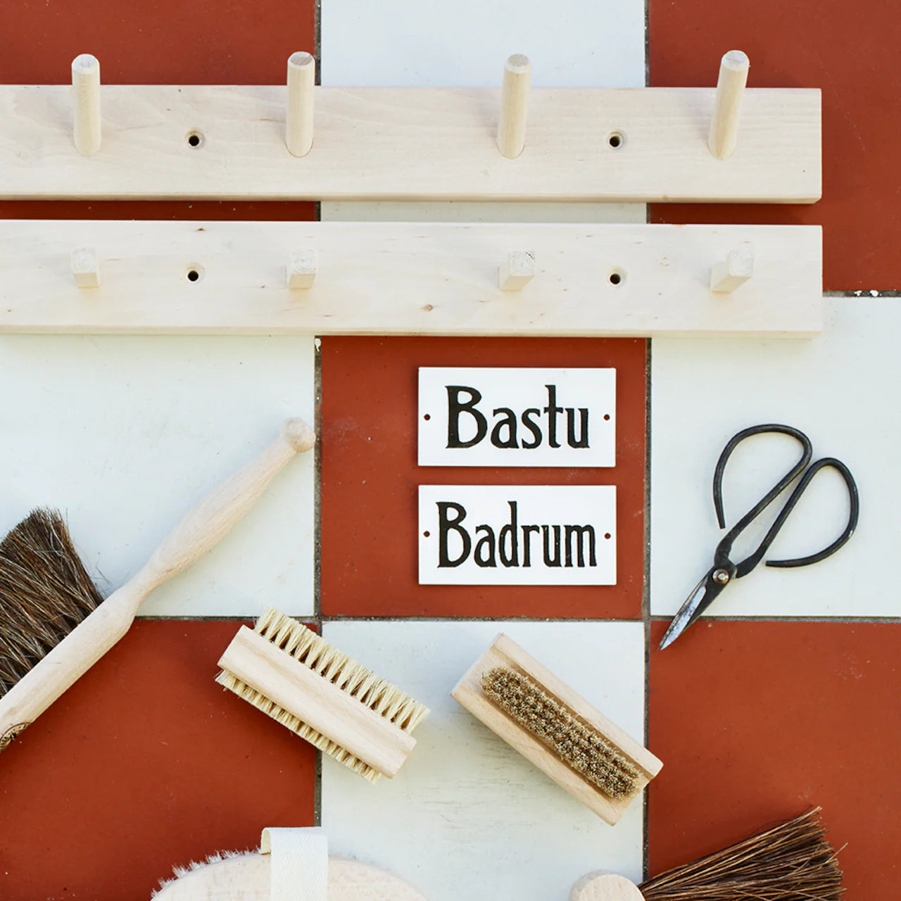 The Strömshaga Nailbrush in Ash Wood, a pair of scissors, two wooden racks, and Bastu and Badrum signs are arranged on a red and white tiled surface.