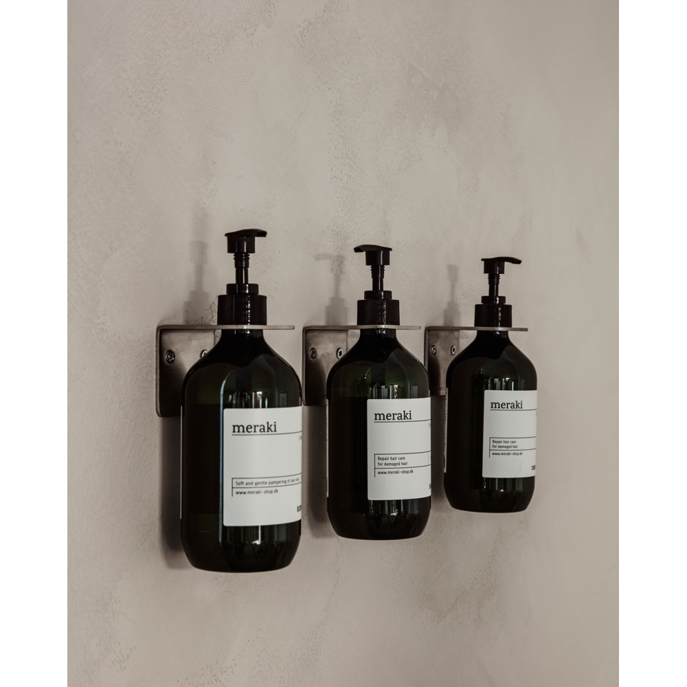 Three Meraki Bottle Hangers in brushed silver finish hold dark green pump bottles with white labels in a neat row on a beige wall, offering stylish and elegant storage for your hand or body care products.