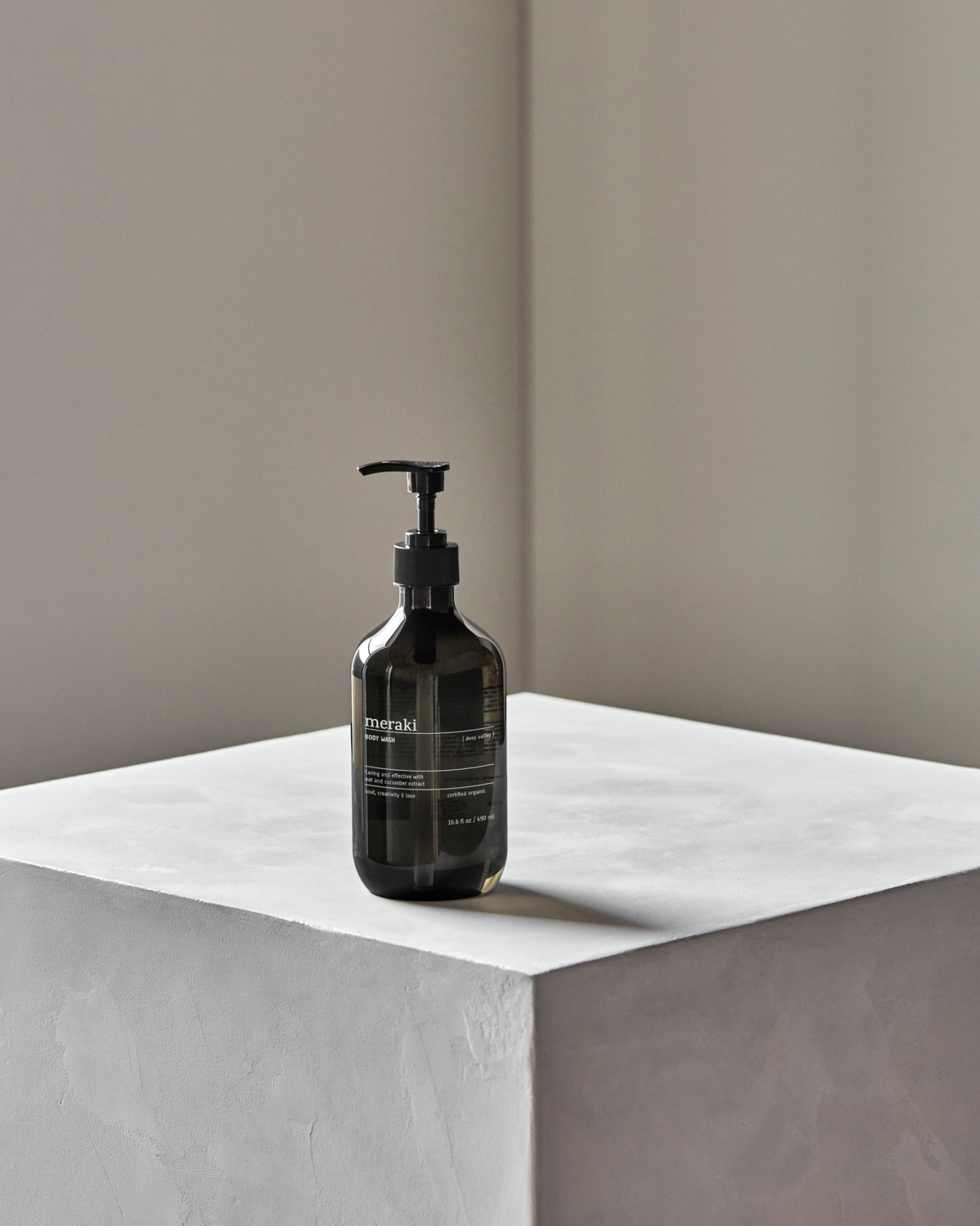 A dark amber Meraki bottle labeled Body Wash, Deep Valley, 490 ml rests on a minimalist light gray pedestal against a plain beige wall.