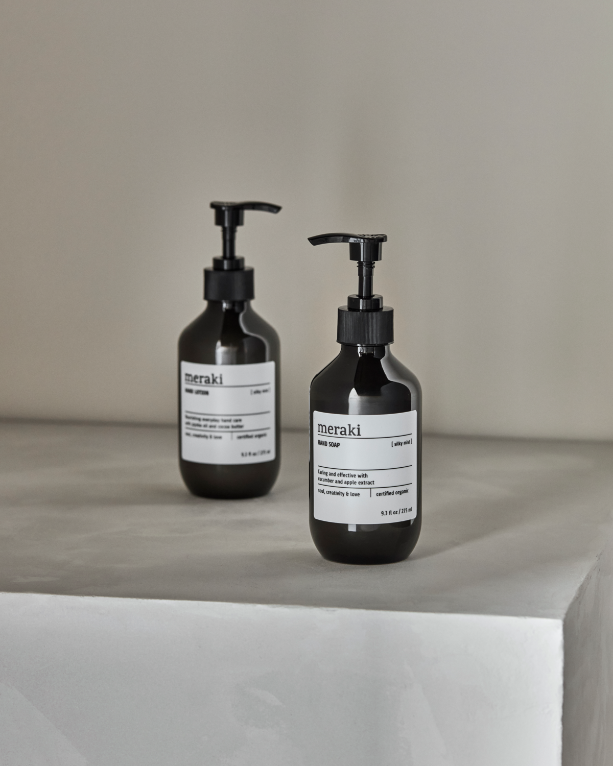 Two black Meraki bottles from the Gift Box Silky Mist Hand Care Set—one hand soap, one hand lotion—are displayed on a light gray minimalist surface, with the lotion bottle in soft focus behind the soap.