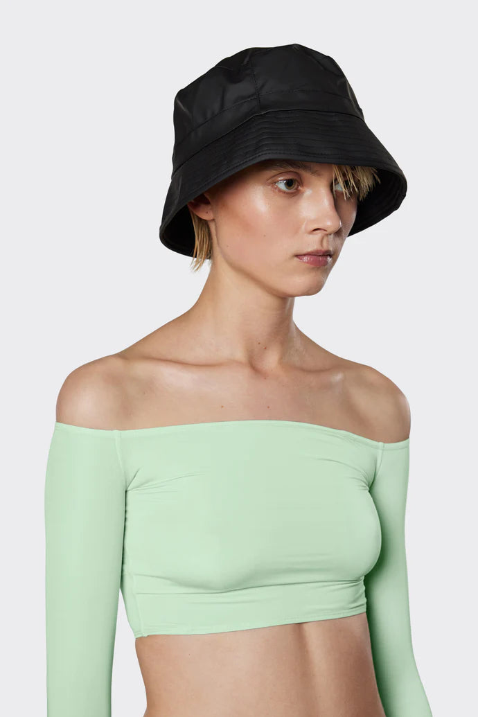 A woman wearing the Rains Bucket Hat in Black by Rains and a light green off-the-shoulder crop top stands against a plain light gray background.