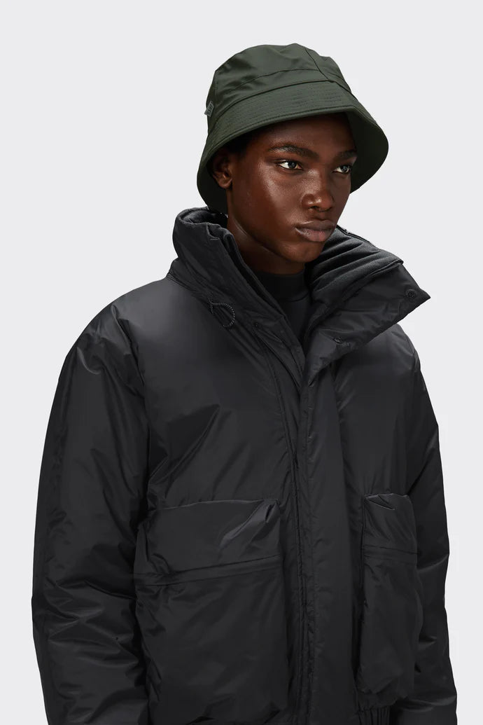 A person wears the Rains Bucket Hat in Green by Rains and a black puffer jacket, standing against a plain light gray background.