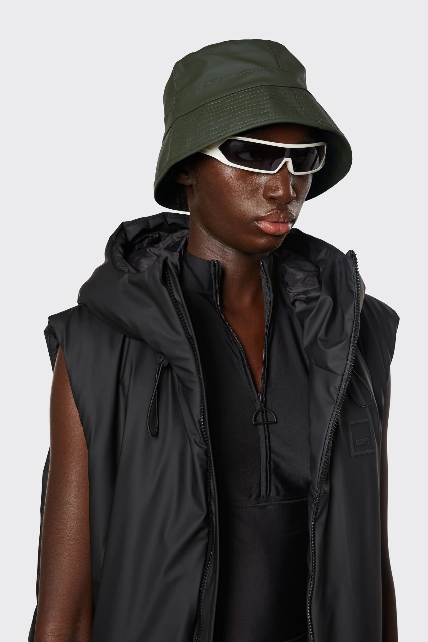 Model wears the Rains Bucket Hat in Green by Rains, paired with white sunglasses, a black zip-up top, and a sleeveless black puffer vest, facing forward against a plain background.
