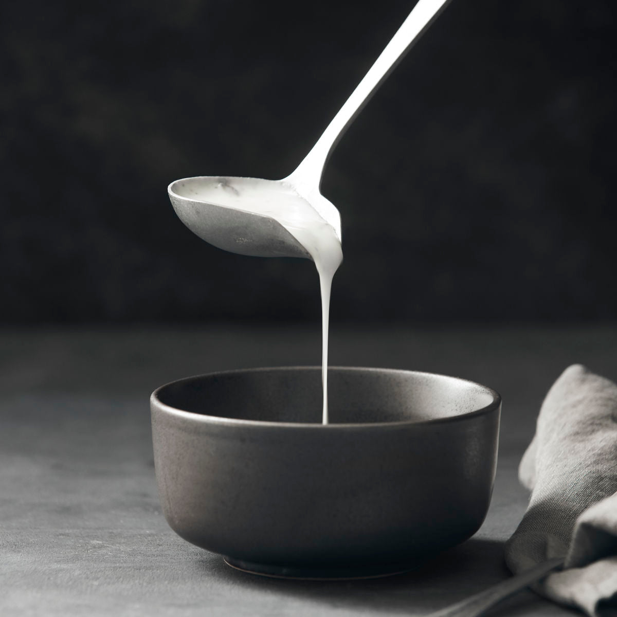 The Nicolas Vahé Daily Sauce Spoon in a silver finish pours a thick white liquid into a matte black bowl, with a folded cloth napkin placed next to the bowl on a gray surface.