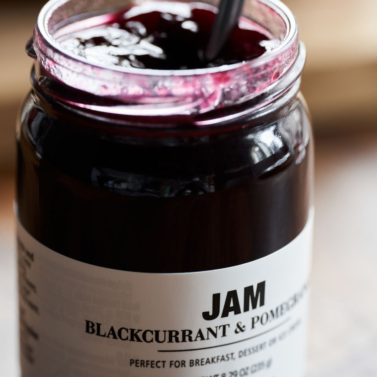 Open jar of Nicolas Vahé Jam - Blackcurrant & Pomegranate with a spoon inside, displaying the dark, glossy fruit spread near the rim of the glass jar.