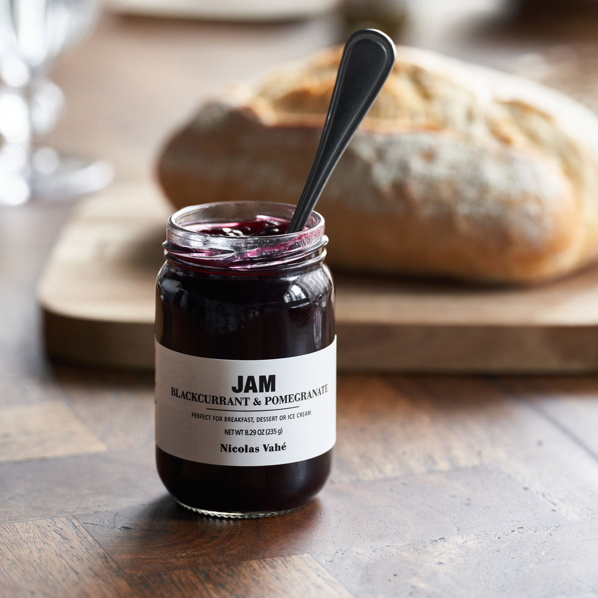 A jar of Nicolas Vahé Jam - Blackcurrant & Pomegranate sits on a wooden surface with a spoon inside, while a loaf of bread rests in the background.