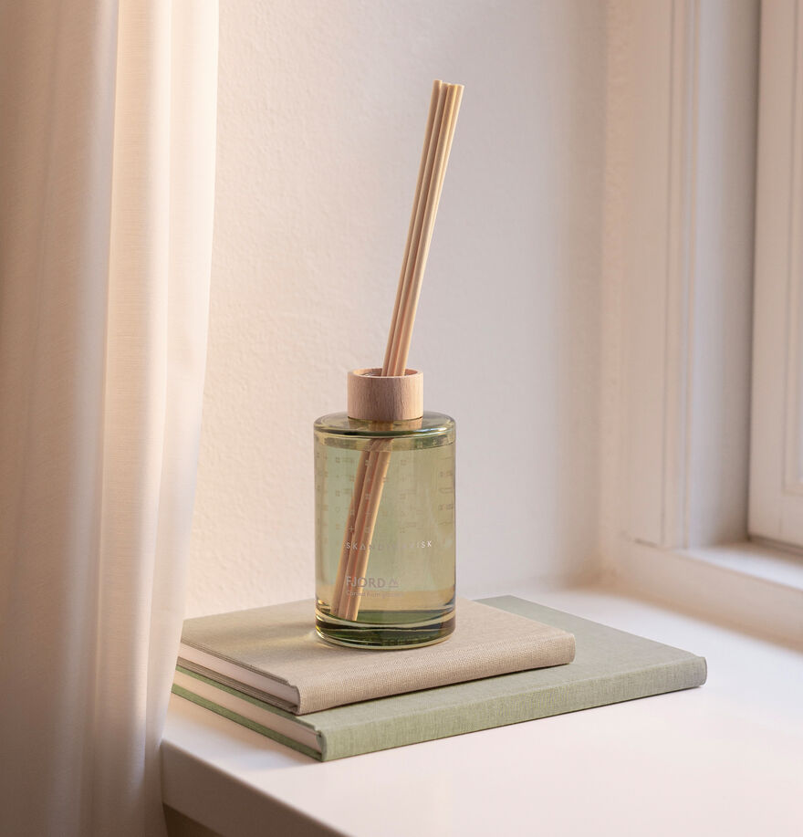 The Skandinavisk Scent Diffuser Fjord 200ml with wooden sticks rests on two closed books by a window and beige curtains, infusing the serene space with fresh vegan fragrance.