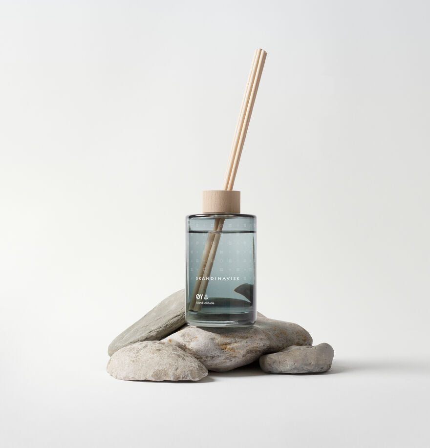 The Skandinavisk Scent Diffuser Øy, 200ml, features clear glass with wooden sticks and cap, displayed on smooth gray stones—a stylish vegan room fragrance for any space.