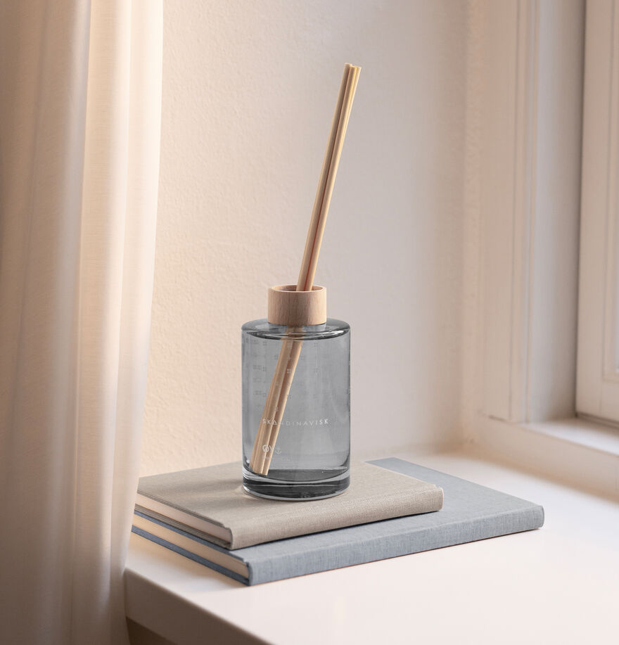 The Skandinavisk Scent Diffuser Øy, 200ml, with two wooden sticks, rests on two closed light-colored books by a window with white curtains, filling the room with a gentle vegan fragrance.