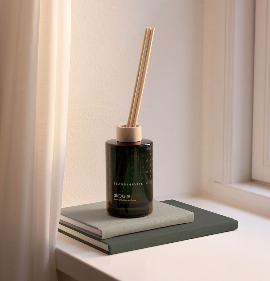 The Skandinavisk Scent Diffuser Skog, 200ml, in a dark glass bottle rests atop two stacked books by a window, sunlight filtering through a white curtain.