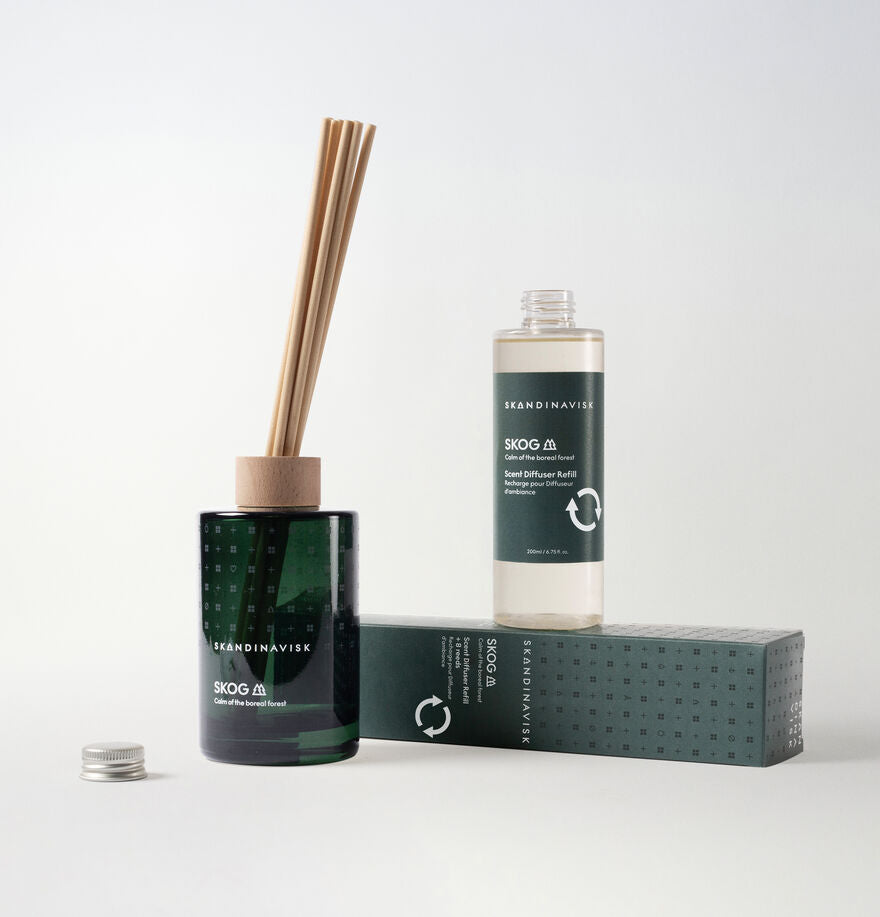 The Skandinavisk Scent Diffuser Skog, 200ml, features a green glass bottle with wooden sticks, a refill, and a box—each labeled by the brand—bringing a Scandinavian fragrance to your space, all set against a white background.