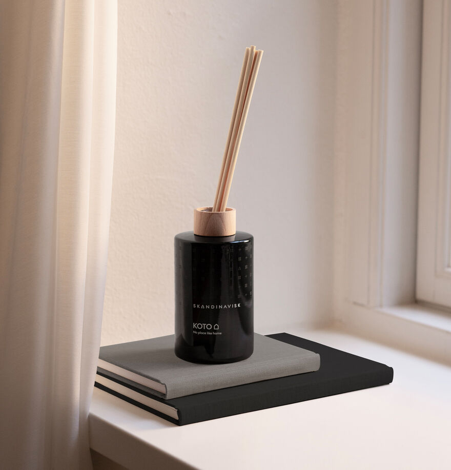 The Skandinavisk Scent Diffuser Koto, 200ml, with wooden sticks sits atop two stacked hardcover books by a window with white curtains, subtly filling the room with a vegan fragrance.