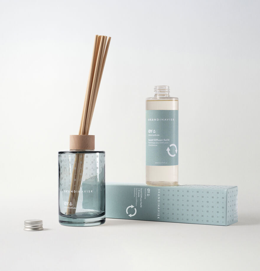 A Skandinavisk Scent Diffuser with reed sticks, a wooden lid, and a light blue label is displayed beside the matching Scent Diffuser Refill Øy (200 ml) and a coordinating rectangular box. All items are set against a plain white background, embodying refined Scandinavian fragrance for any room.