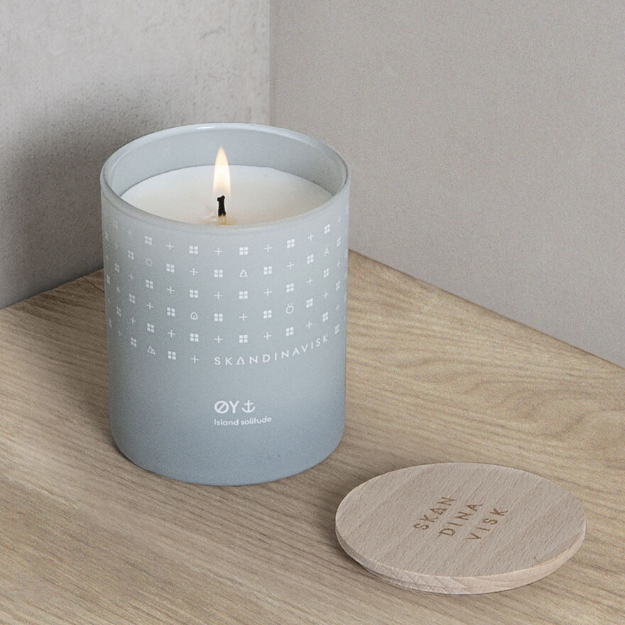 The Skandinavisk Scented Candle Oy 50h, 200g glows in a light gray container on a wooden surface, its white Swedish rapeseed wax filling the room with scent beside the matching lid.