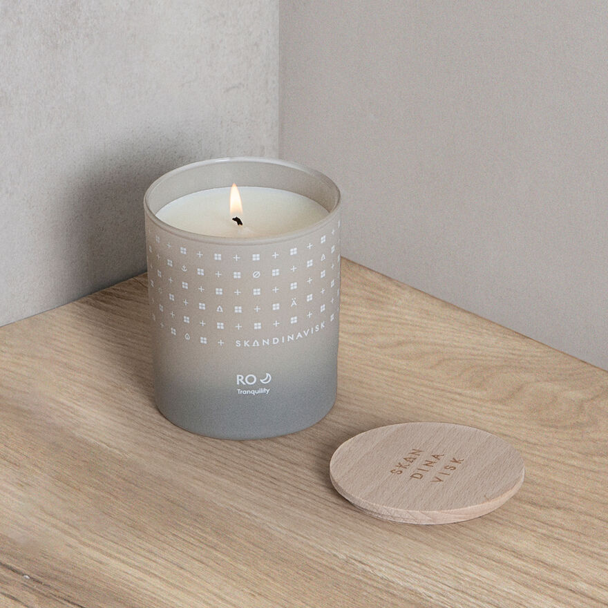 A Skandinavisk Scented Candle Ro 50h, 200g in frosted glass with Swedish rapeseed wax rests on a wooden surface beside its round wooden lid.