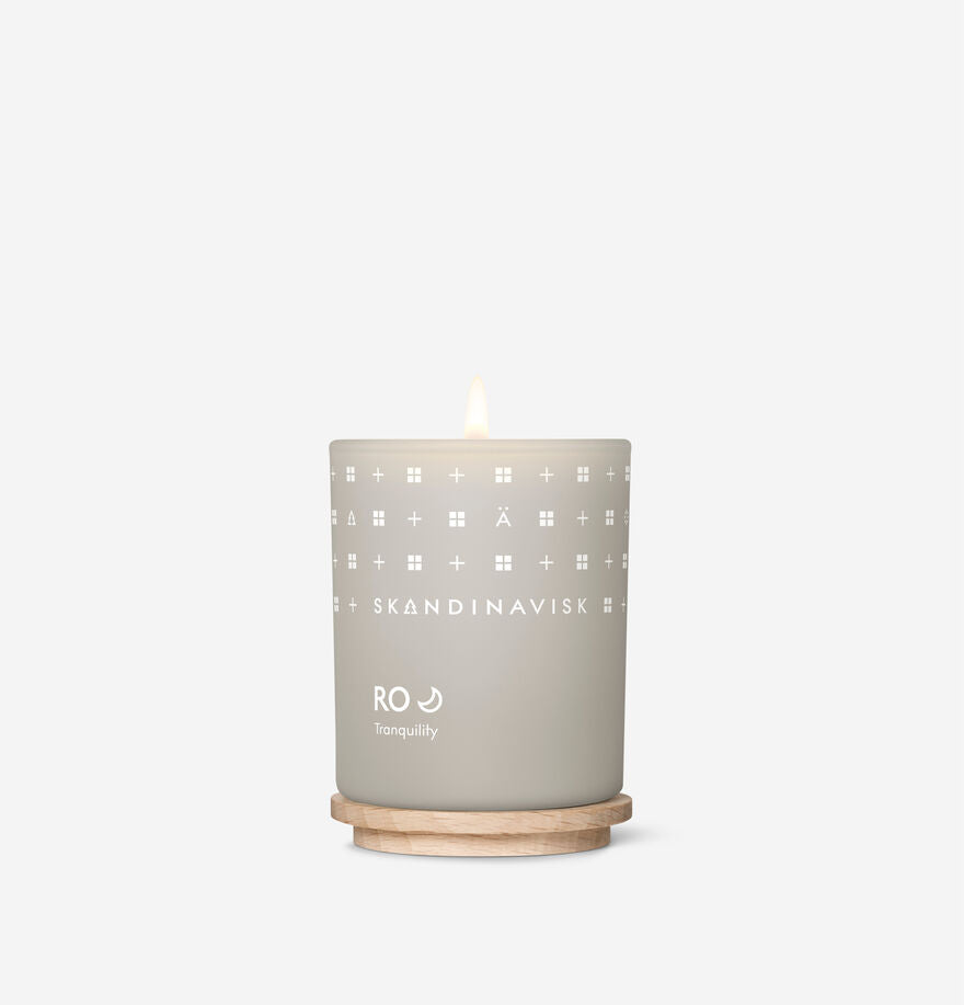 The Skandinavisk Mini Scented Candle Ro 20h, 65g, in a light grey glass holder with white geometric patterns and made from Swedish rapeseed wax, rests on a wooden coaster against a white background.