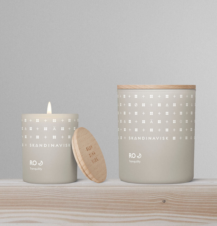 Two beige Skandinavisk Mini Scented Candle Ro 20h, 65g candles sit on a wooden surface—one lit, the other with its lid beside it. Made from Swedish rapeseed wax, they bring a calming ambiance to any space.