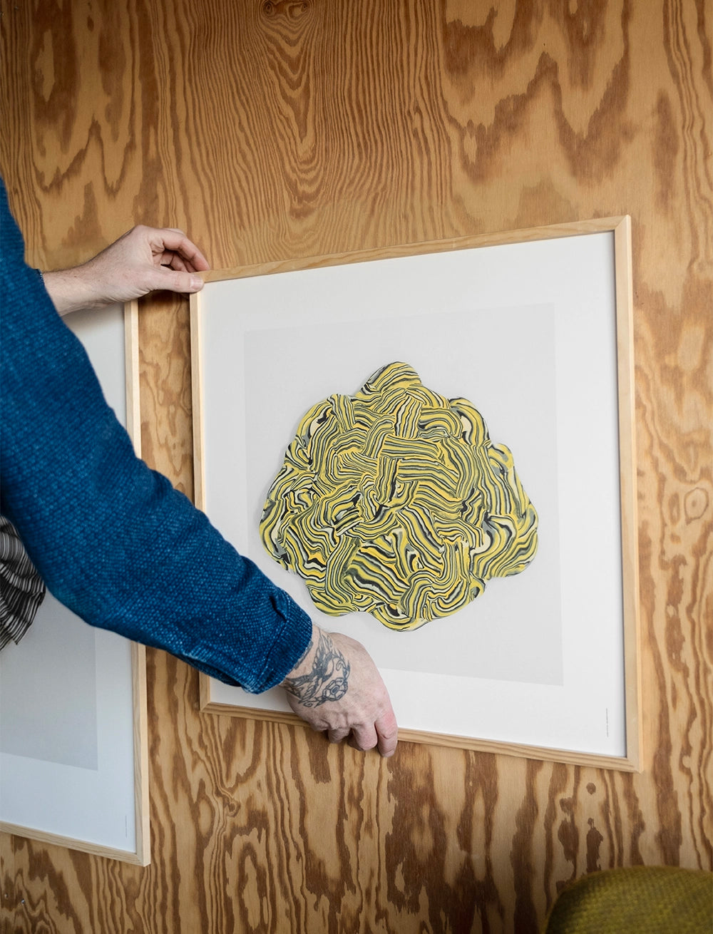 A person hangs the Fine Little Day Vibrant Print (50 cm x 50 cm), an abstract artwork with yellow and black swirls, on a plywood wall, bringing poetic energy to the space.
