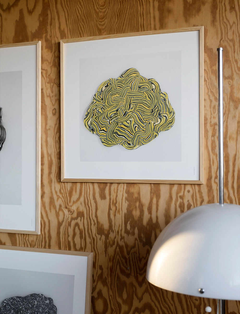 The Vibrant Print (50 cm x 50 cm) by Fine Little Day, featuring yellow and black lines, hangs on a wood-paneled wall next to a metallic lamp and other framed art, adding poetic energy to the space.