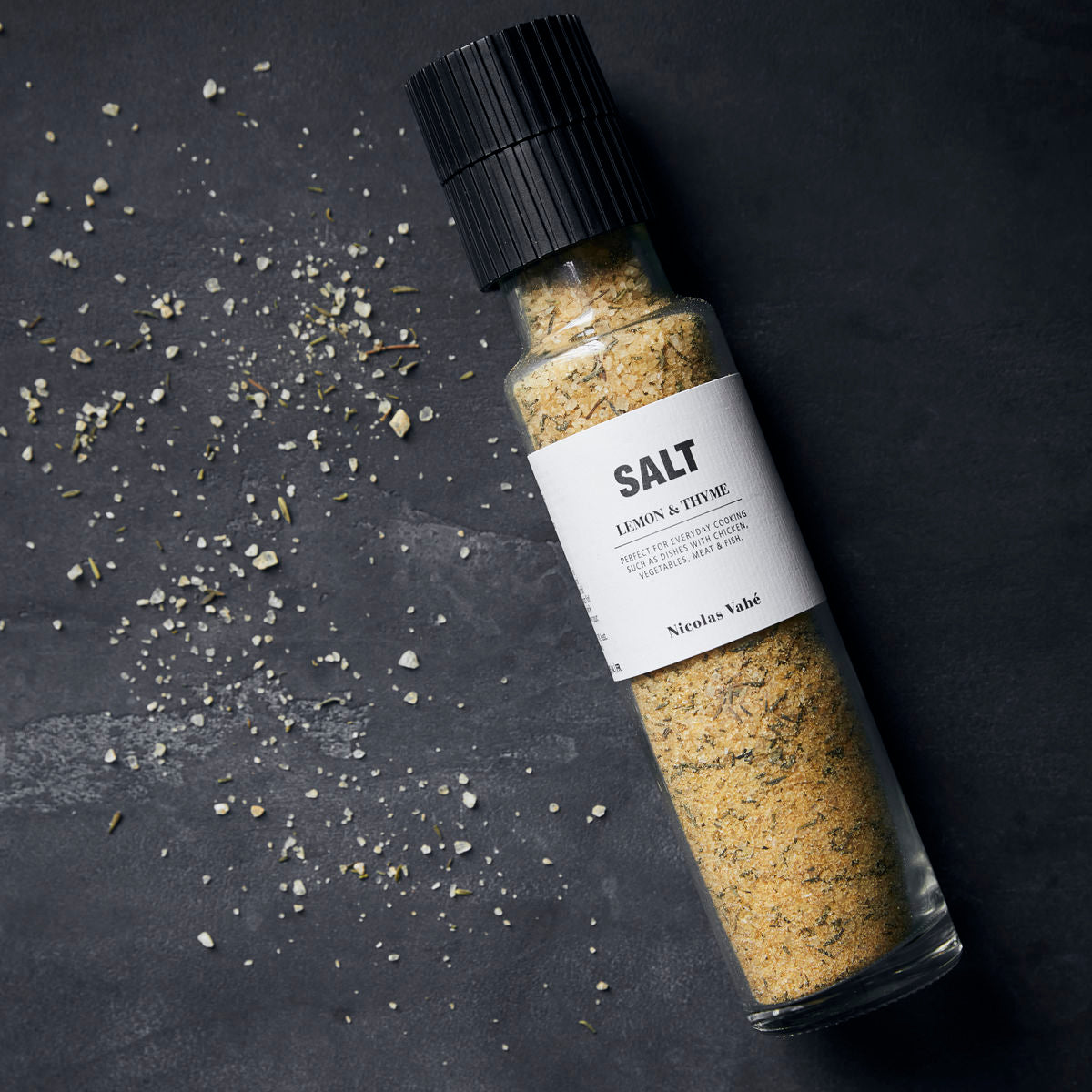 A Nicolas Vahé Salt - Lemon & Thyme glass grinder bottle with a ceramic top rests on a dark surface, its coarse sea salt blend scattered beside it.
