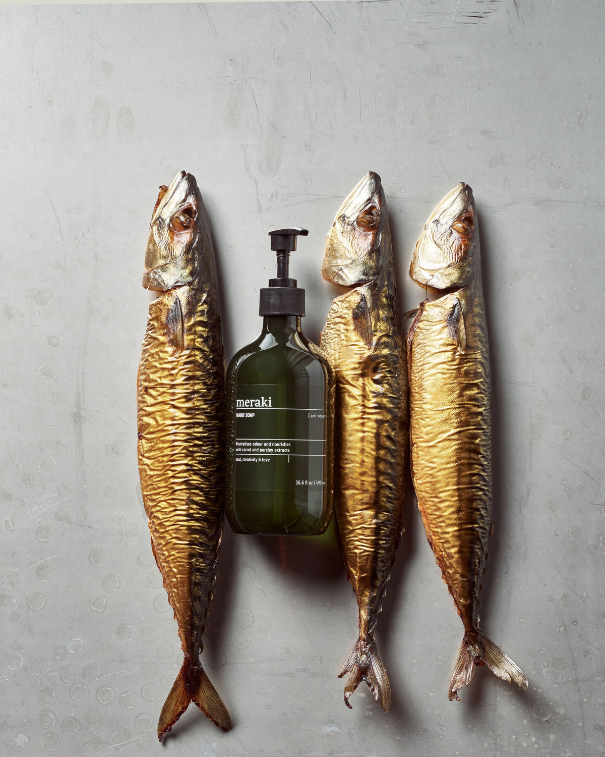 Three smoked fish lay side by side on a gray surface, with a bottle of Meraki Hand Soap, Anti-Odour—designed to neutralise food smells—positioned between the second and third fish.