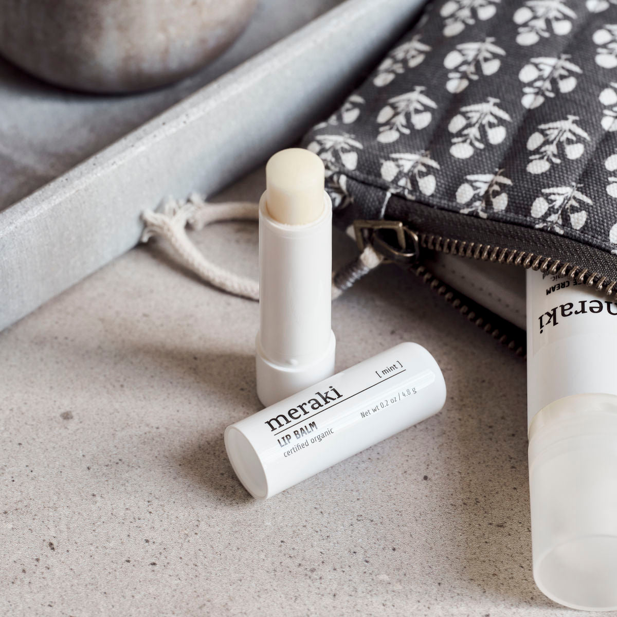 An open white tube of Meraki Organic Lip balm, Mint is on a counter beside a gray pouch and another Meraki product lying on its side.