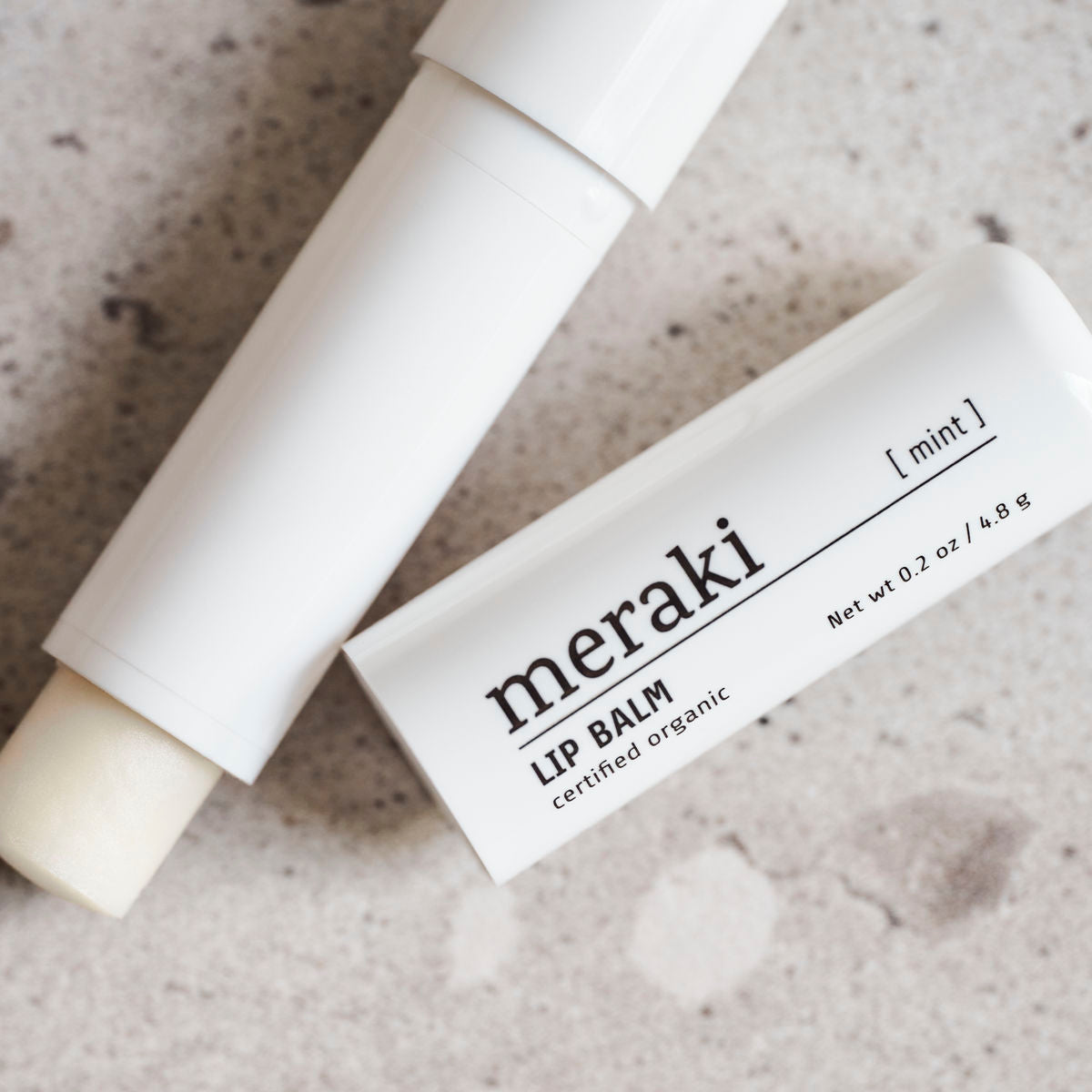 An open white tube of Meraki Organic Lip Balm, Mint, rests on a speckled surface with its cap beside it.