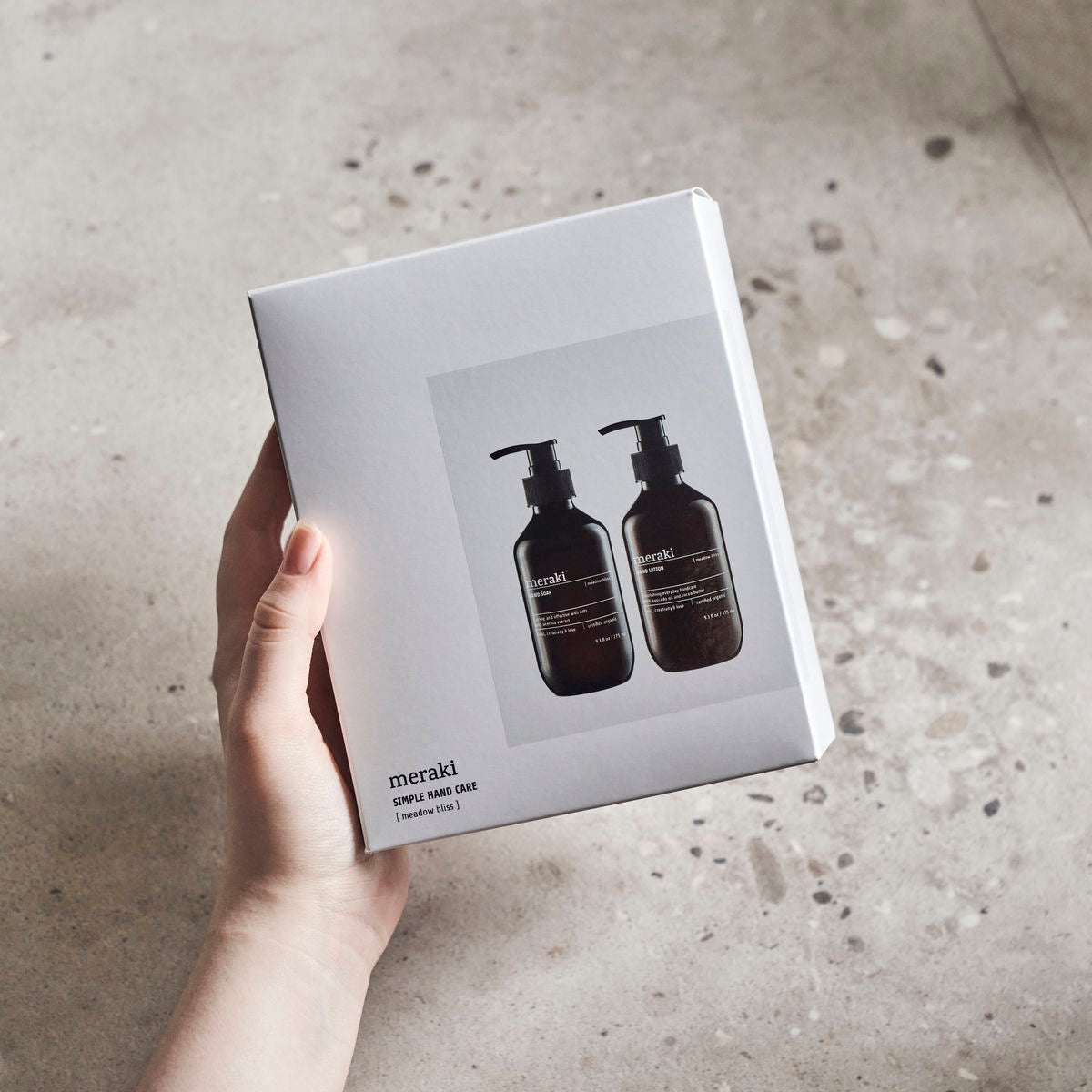 A hand holds a white gift box featuring Meraki’s “Giftbox Meadow Bliss Organic” hand soap and lotion, both in dark pump bottles, set against a speckled neutral background.