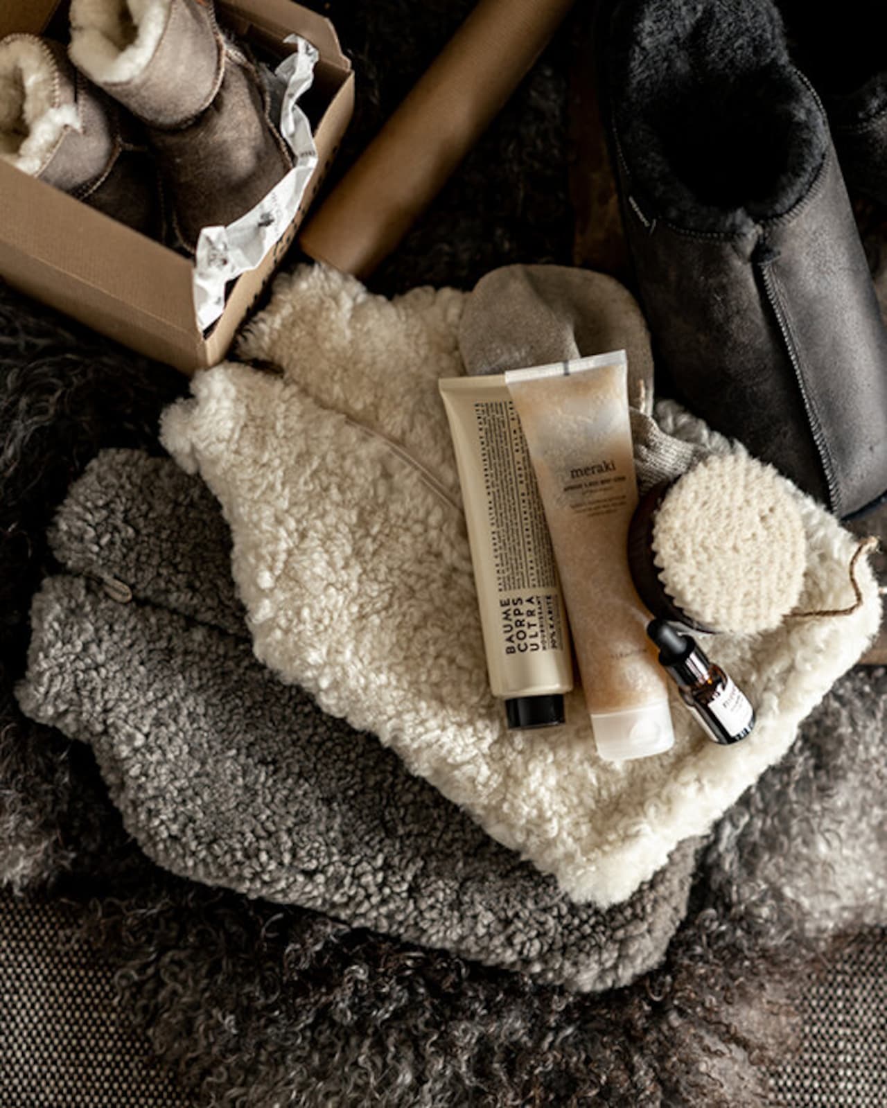 Shepherd of Sweden’s Kerri sheepskin hot water bottle cover in Carbon is styled with slippers, a fluffy throw, skincare, and a body brush on textured wool—perfect for cozy winter muscle relief.