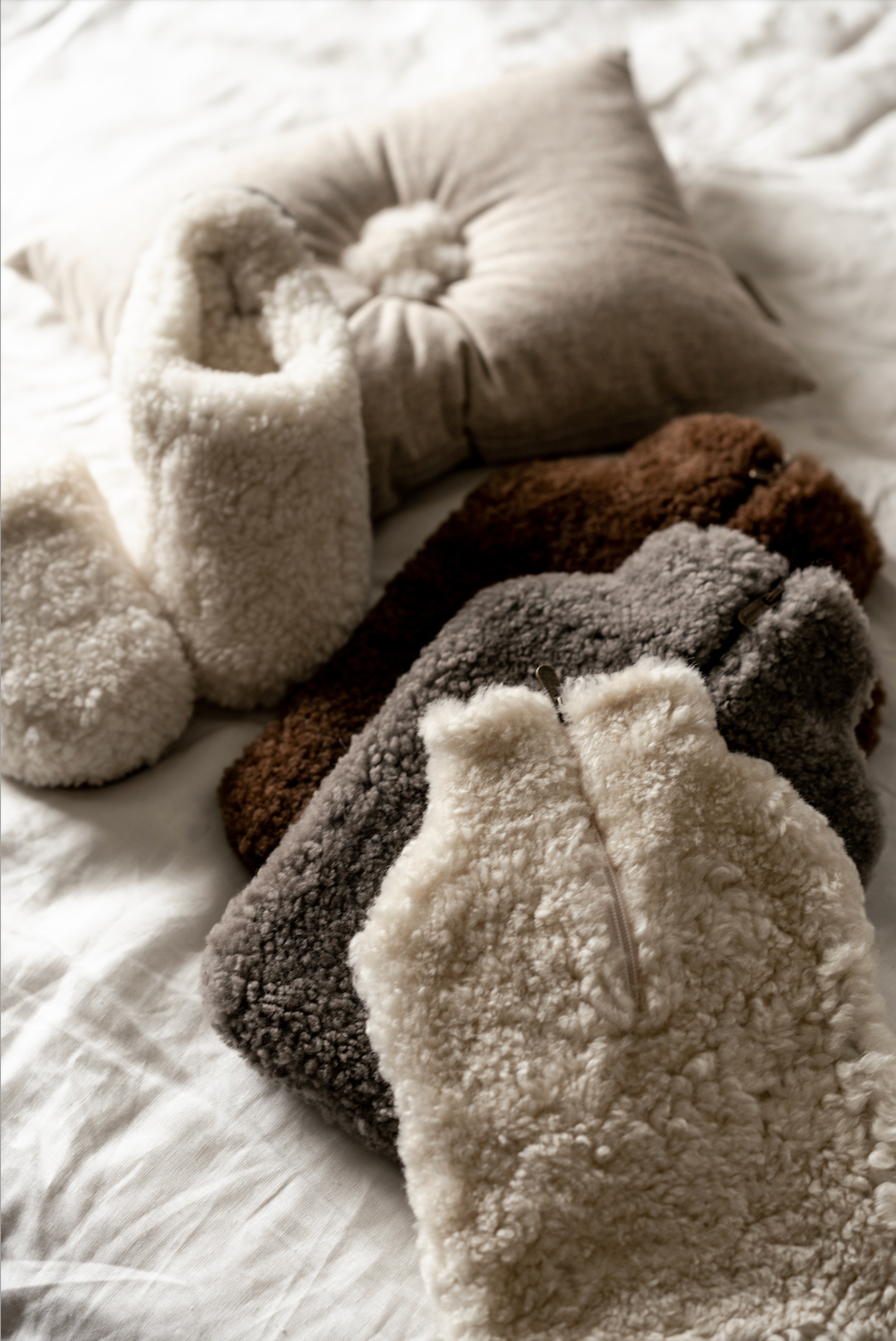 Three sherpa vests, a pair of cream sherpa slippers, a beige cushion, and the Shepherd of Sweden Kerri Hot Water Bottle Cover in Stone sheepskin are arranged on a white bedspread.