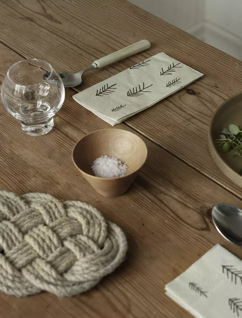 A wooden table set with a glass, a wooden salt bowl, cutlery, rope trivet, and Fine Little Day’s Pine (Tall) Paper Napkins (33 x 33 cm) creates inviting settings with their simple Scandinavian leaf pattern.