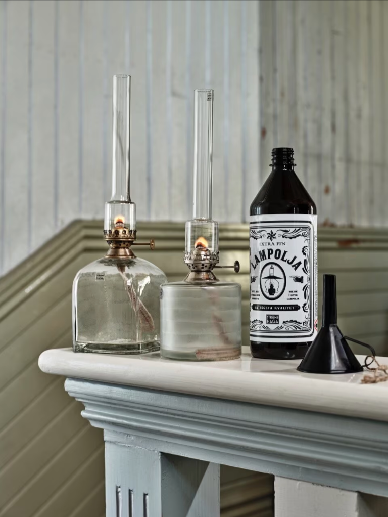 Two Strömshaga Kerosene Lamps Elisabeth in glass with antique brass burners and flames stand on a table next to a black Lampolja bottle and a small funnel, all set against paneled walls. Each lamp measures 33cm and features a square design.