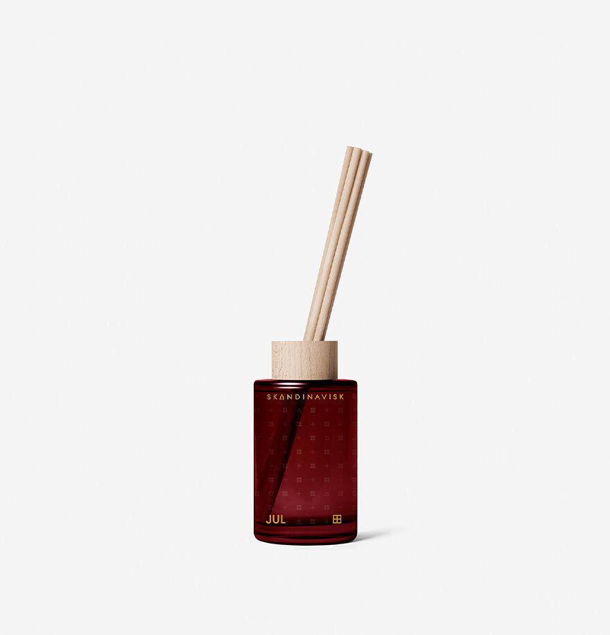 The Skandinavisk Jul Scent Diffuser, 100 ml, features a red glass bottle with a wooden cap and two reeds, capturing a Scandinavian Christmas with its spiced orange fragrance, presented on a simple white background.