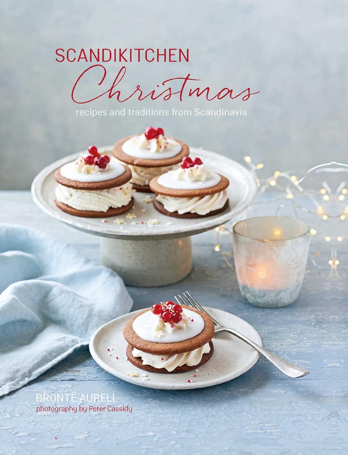 The book ScandiKitchen Christmas – Recipes and Traditions From Scandinavia by SkandiKitchen features festive recipes, pictured with four sandwich cookies topped with white icing and red berries, plus a lit candle and blue napkin.