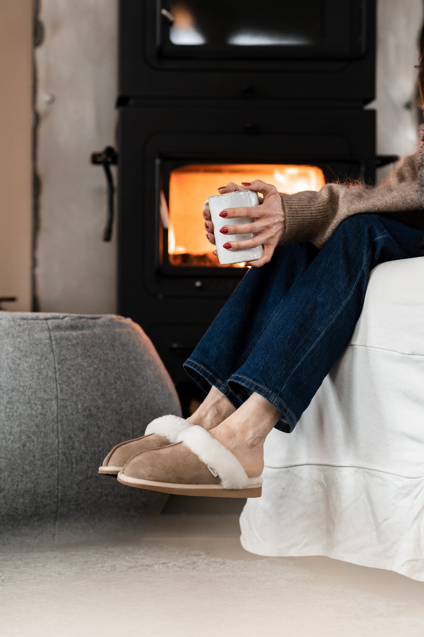 A person in Shepherd of Sweden's Jessica Sheepskin Slippers in Chestnut and jeans relaxes on a white sofa, holding a mug. A wood-burning stove with visible fire glows in the background, creating a cozy indoor vibe.