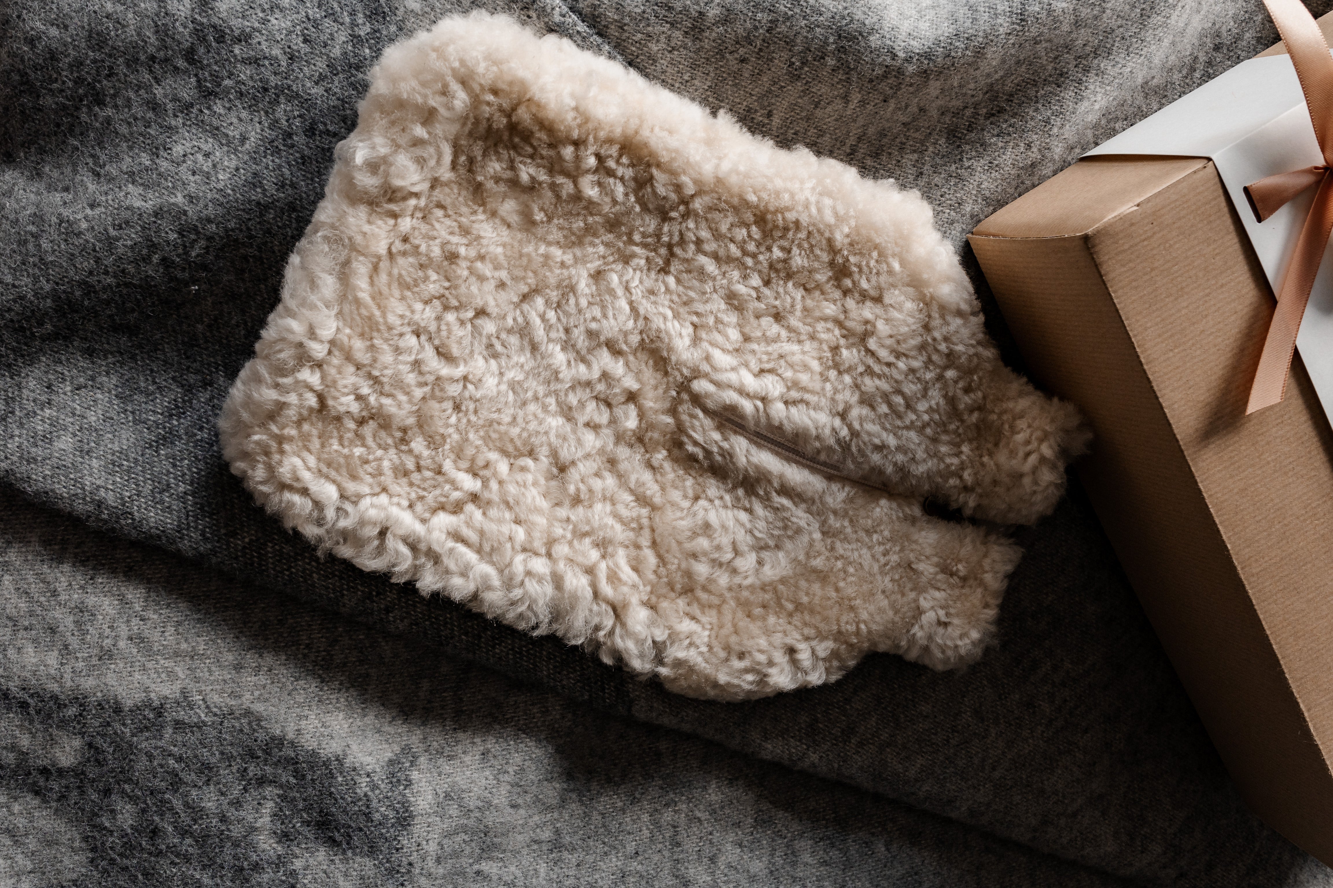 The Kerri Hot Water Bottle Cover in Cream Sheepskin by Shepherd of Sweden rests on a grey wool blanket beside a brown gift box with a pink ribbon—perfect for soothing muscles and cozy nights.