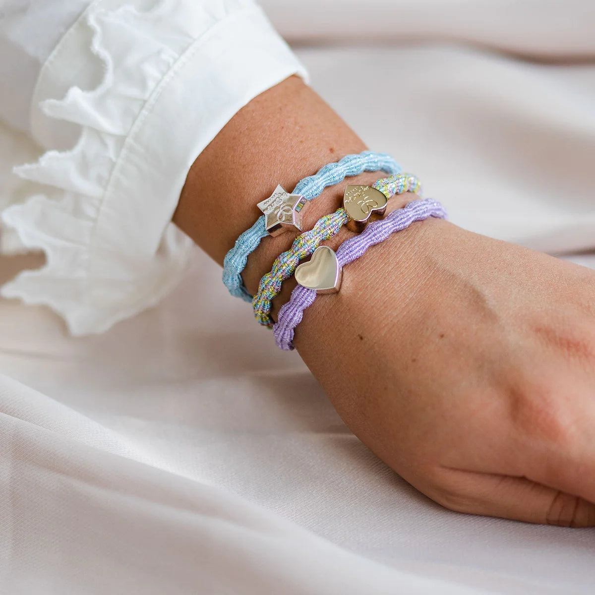 A hand wearing a white ruffled sleeve displays three colorful ByEloise Bangle Bands from the Sparkle Hair Accessory Collection, each crafted with metallic thread elastic and adorned with a different metallic charm—including a gold heart, star, and round disc. The soft, light-toned fabric background enhances the delicate appeal of these Candy collection accessories.