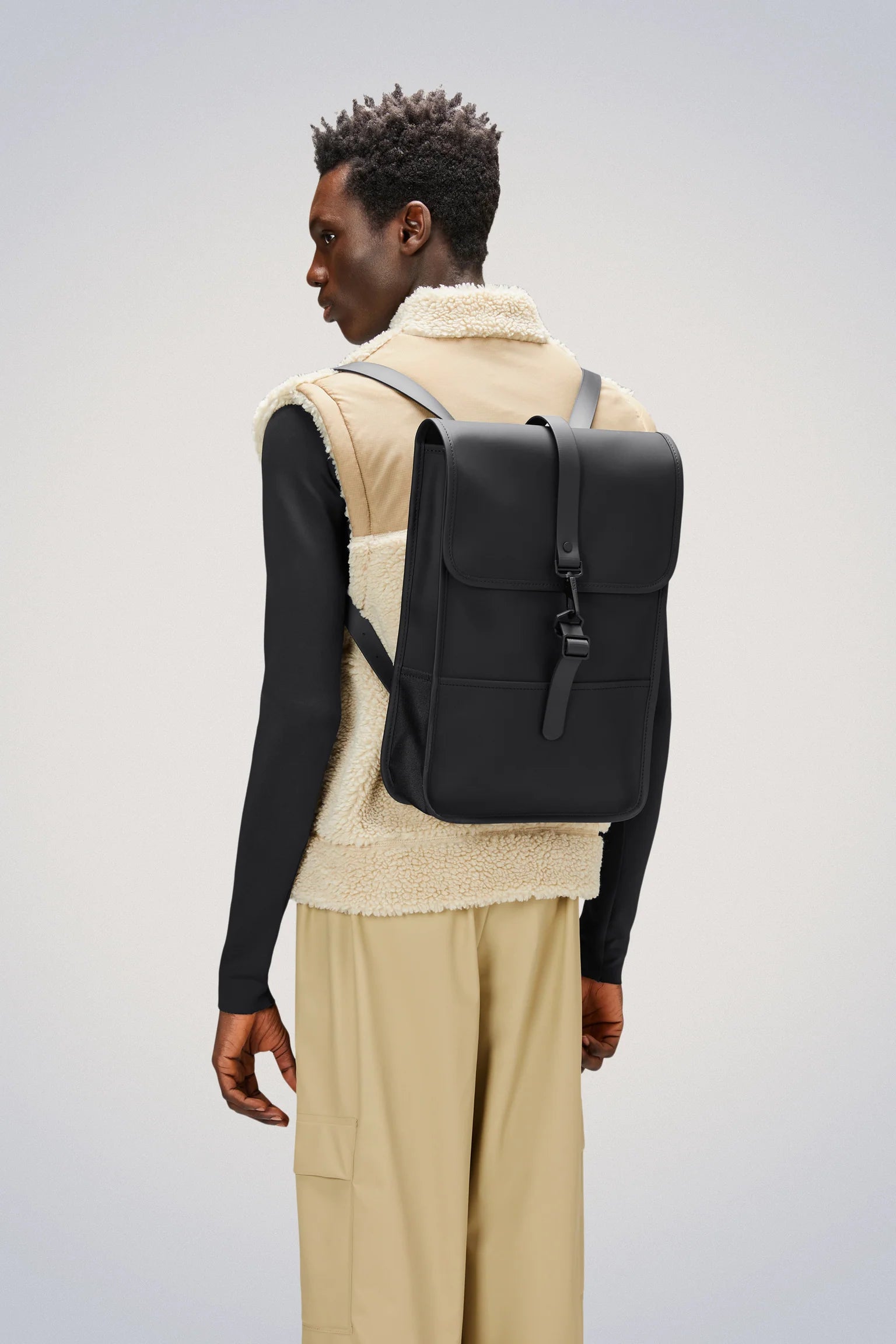 A person, dressed in a beige fleece vest over a black long-sleeve shirt and tan pants, stands sideways against a plain light background, carrying the Rains Backpack Mini Bag in Black by Rains on their back.