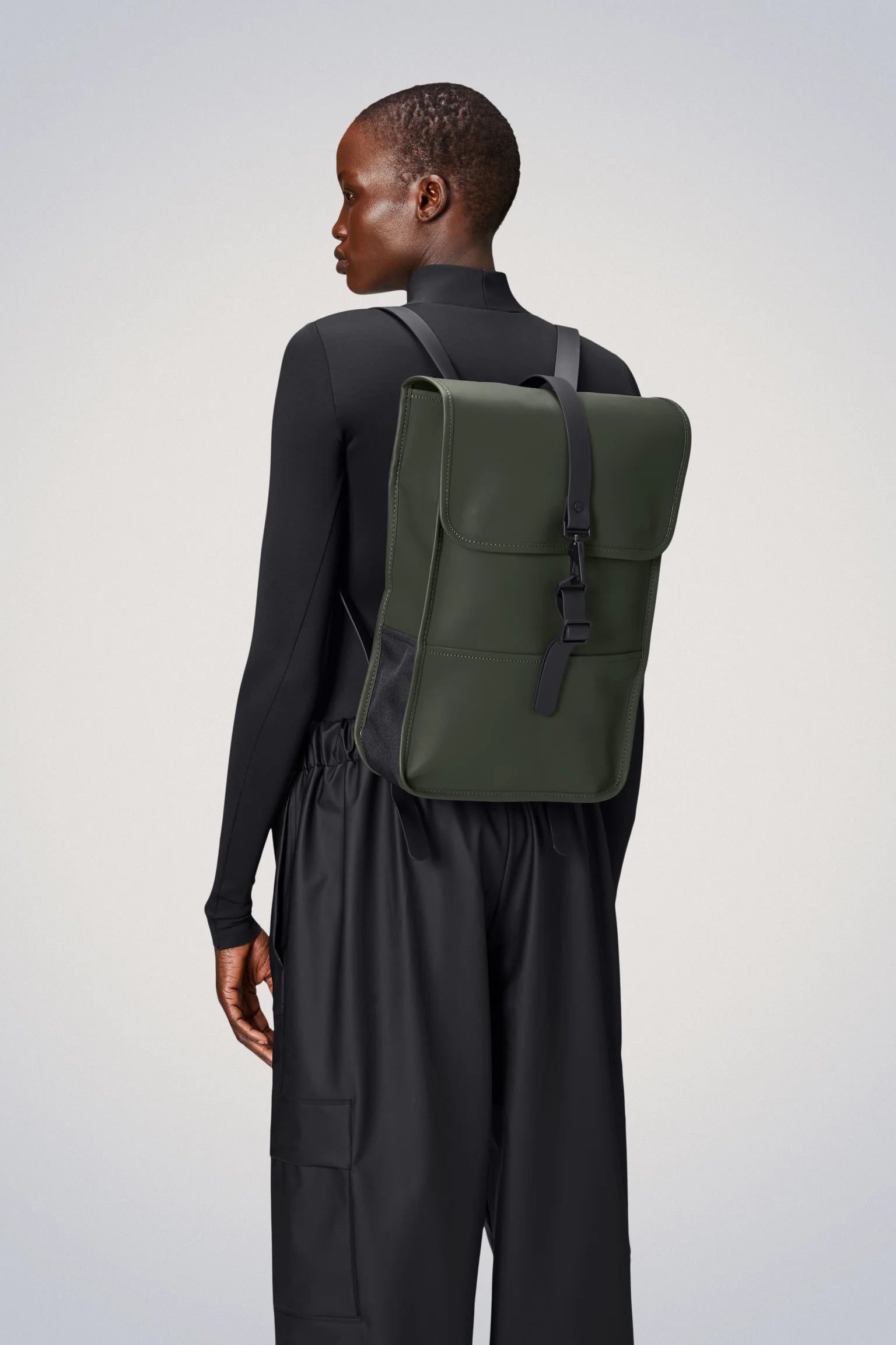 A person wearing a black long-sleeve top and black pants, shown from the back, is carrying the Rains Backpack Mini Bag in Green by Rains—a sleek, rectangular laptop backpack featuring a buckle closure.