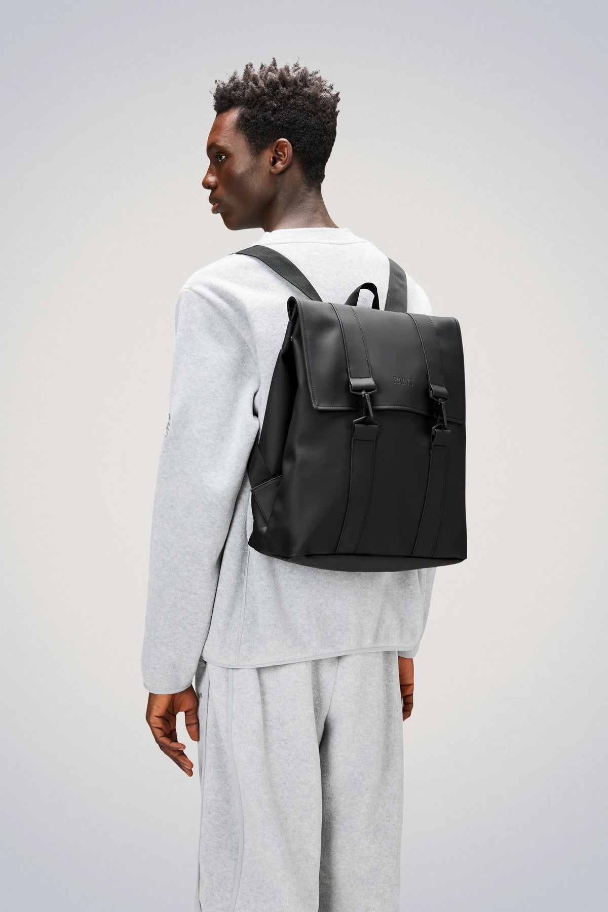 A person wearing a light gray sweatshirt and sweatpants stands sideways, looking back. They have a short Afro hairstyle and are carrying the Rains MSN Bag in Black by Rains—a sleek commuter bag with two buckle straps and a convenient laptop pocket.