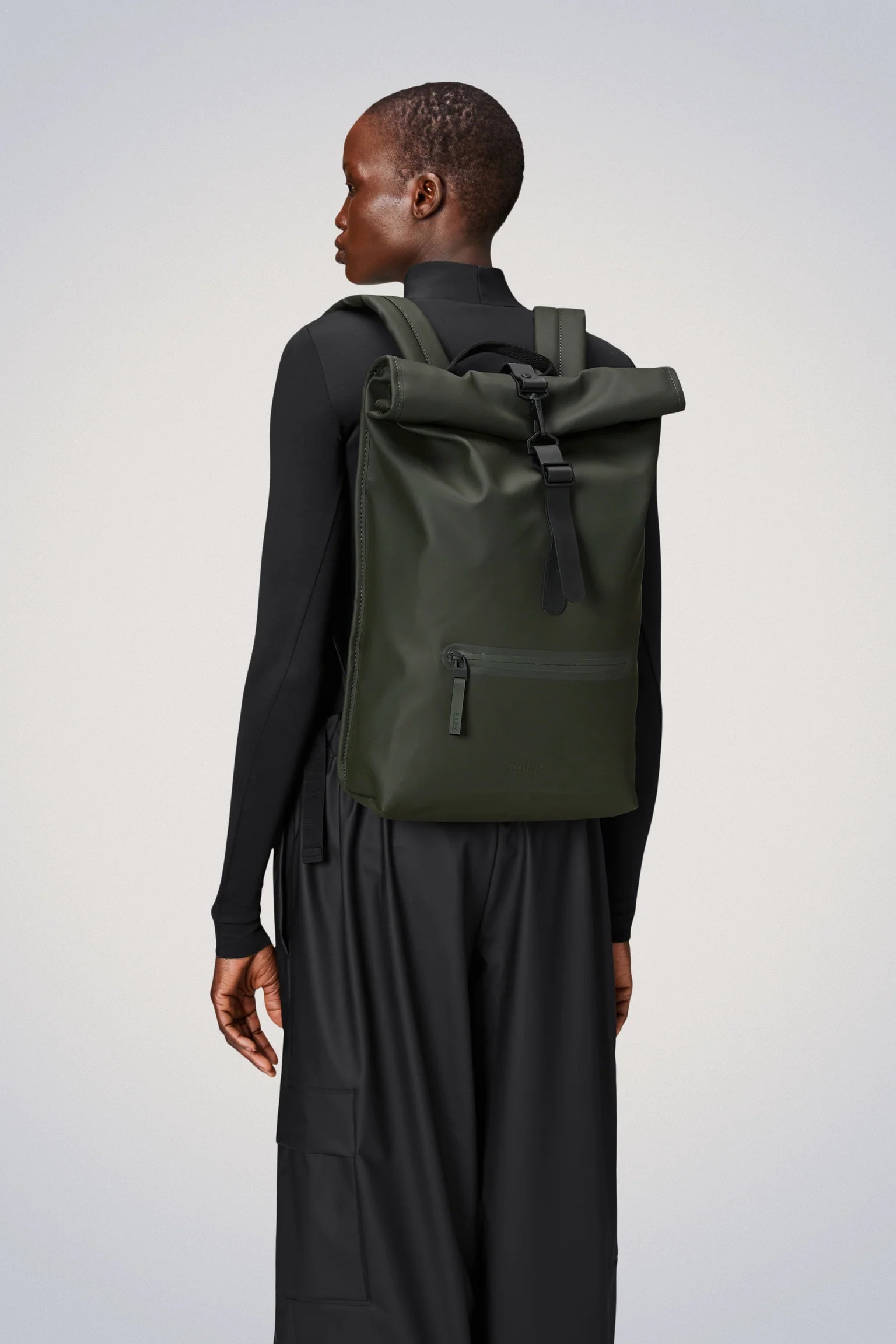A person wearing the Rains Rolltop Rucksack Backpack in Green by Rains, along with a black long-sleeve top and black pants, stands against a plain light gray background.