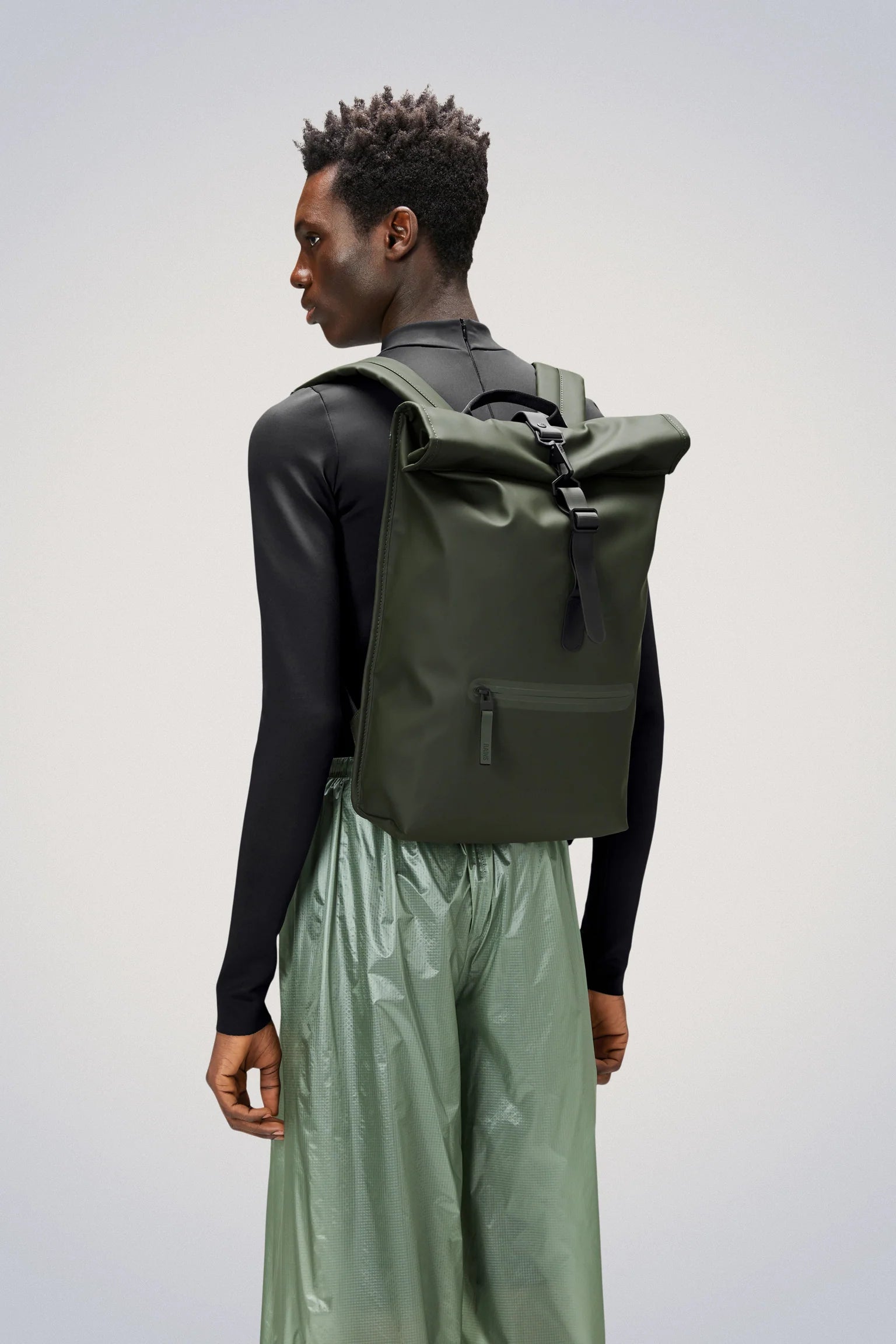 A person wearing the Rains Rolltop Rucksack Backpack in Green over a black long-sleeve shirt and light green pants stands against a plain background.