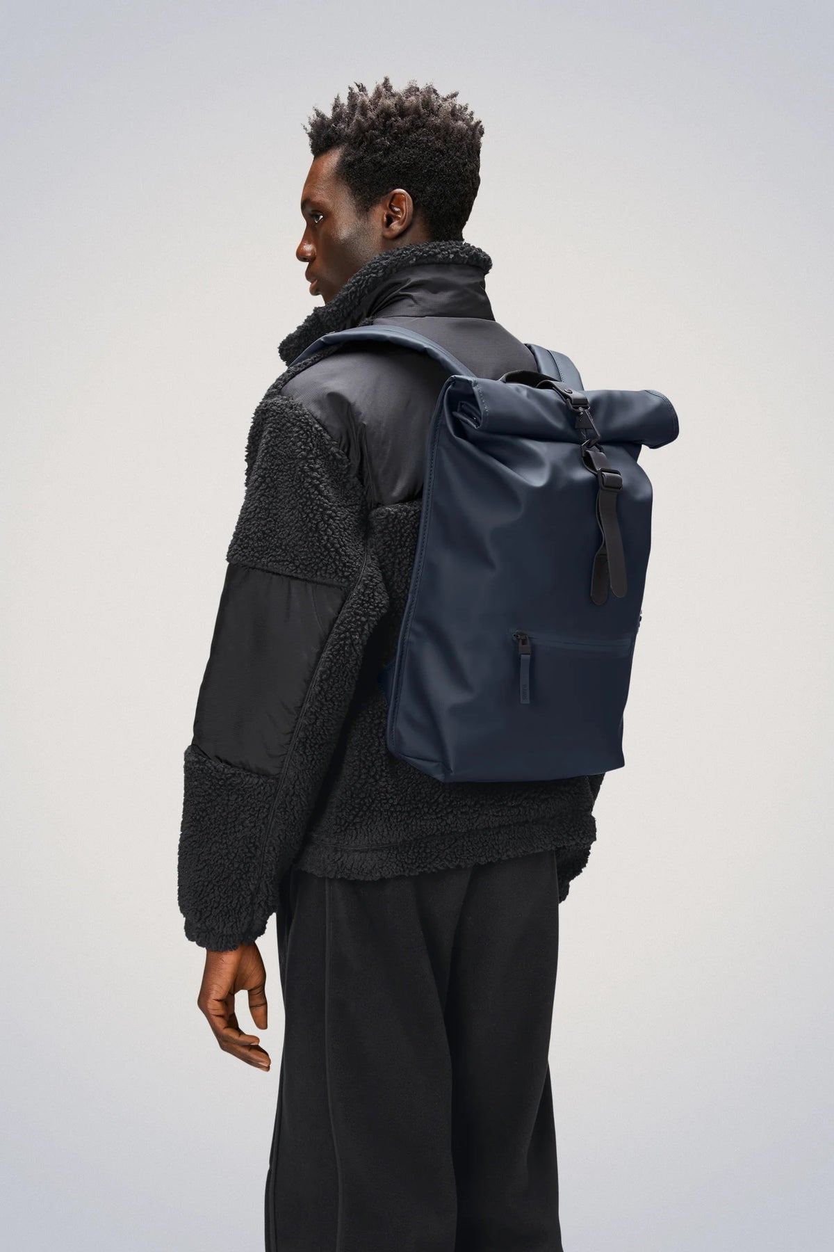 A person dressed in a black jacket and black pants is shown from the side, carrying the Rains Rolltop Rucksack Backpack in Navy by Rains over one shoulder against a plain light background.