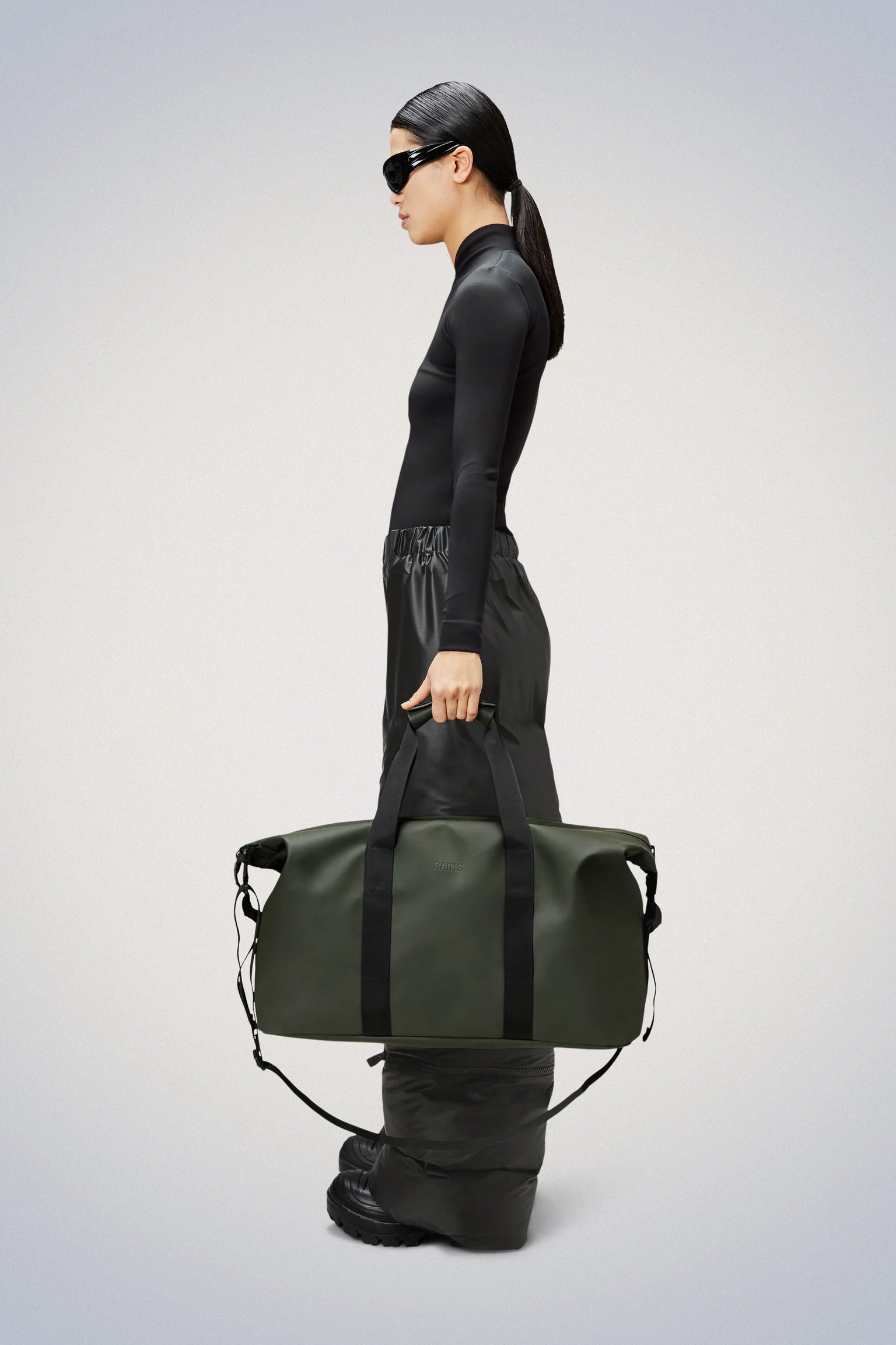 A person in black clothes and sunglasses stands in profile, holding the Rains Hilo Weekend Bag in Green—a spacious waterproof travel bag from Rains—against a plain background.