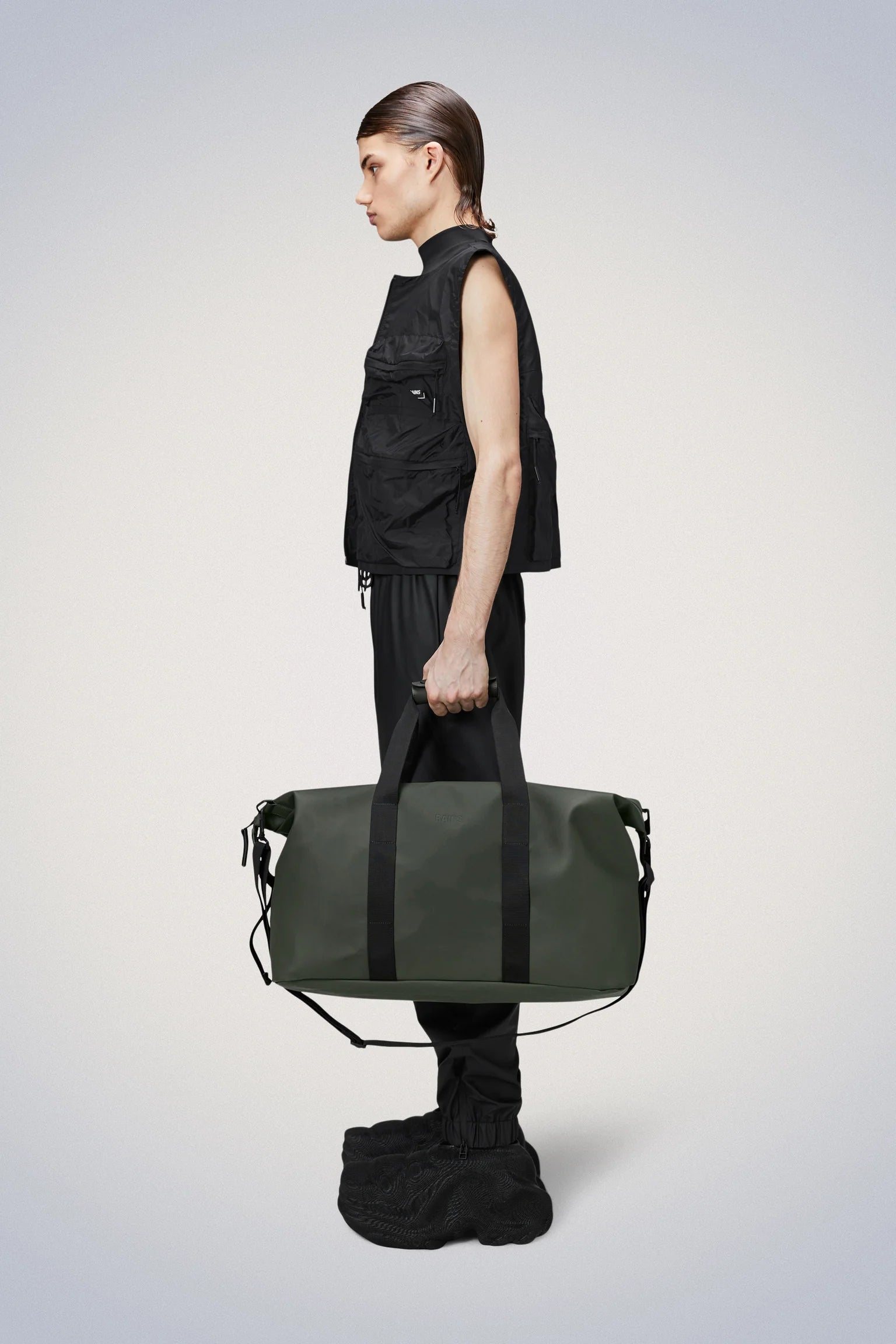 A person in black clothing and large black shoes holds the Rains Hilo Weekend Bag in green, a waterproof PU travel bag by Rains, against a plain background.