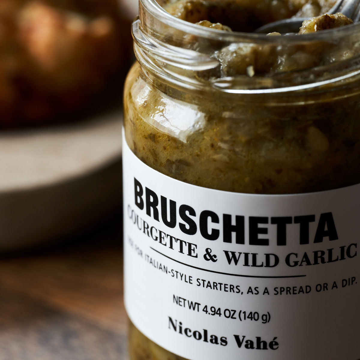 A jar of Nicolas Vahé Bruschetta, Courgette & Wild Garlic sits on a wooden surface, its vibrant spread visible through the glass. In the background, a bruschetta is softly blurred.