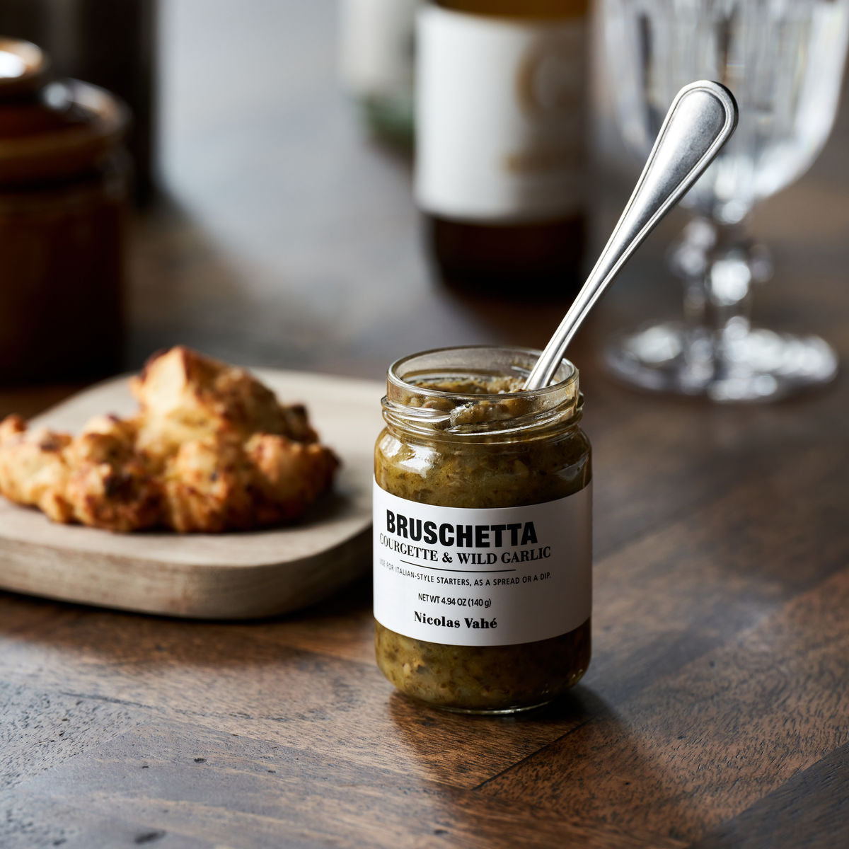 A jar of Nicolas Vahé Bruschetta, Courgette & Wild Garlic with a spoon sits on a wooden table beside a plate of two bread slices; glasses and bottles are blurred in the background.