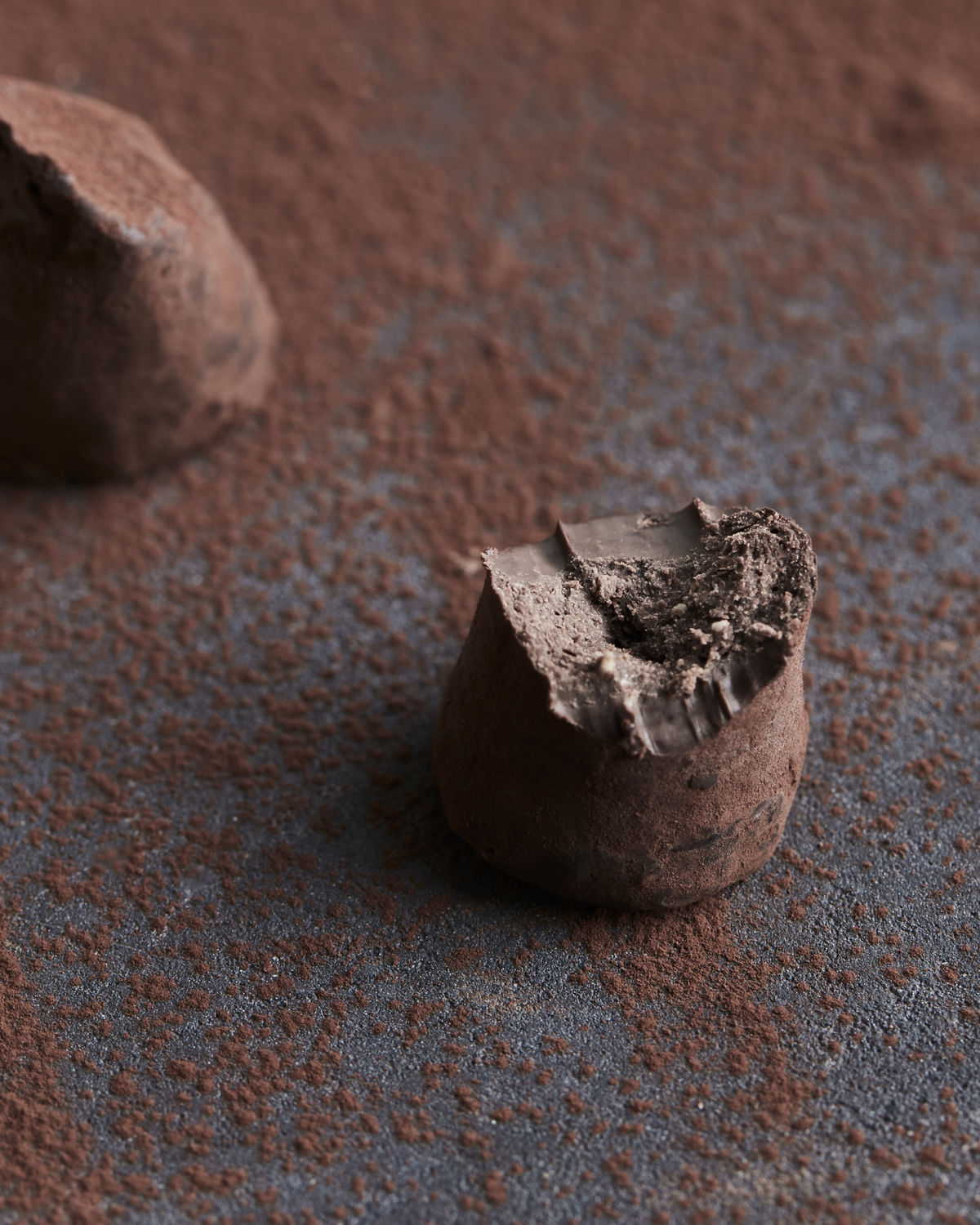 A close-up of a bitten Chocolate Truffles, Pistachio & Crunch by Nicolas Vahé, dusted with cocoa powder and resting on a textured surface with scattered cocoa—an artisanal delight.