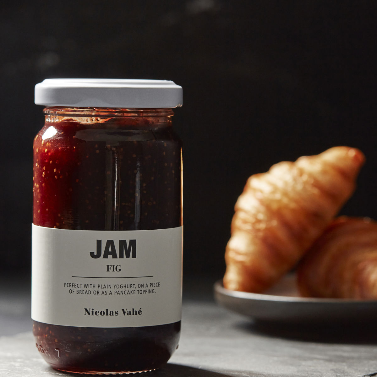 A jar of Nicolas Vahé Jam, Fig sits on a surface, ideal for pairing with cheese, while two croissants rest on a black plate in the blurred background.