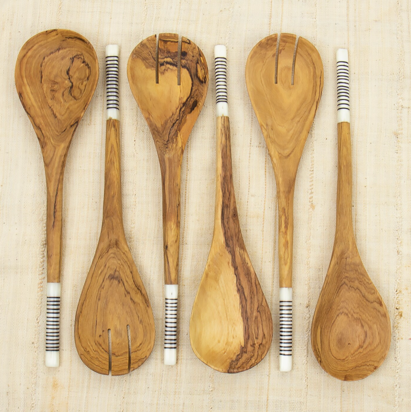 Five Striped Wooden Salad Servers by A World of Craft from Kenya, with black and white striped handles, are arranged in a row on a light fabric surface.